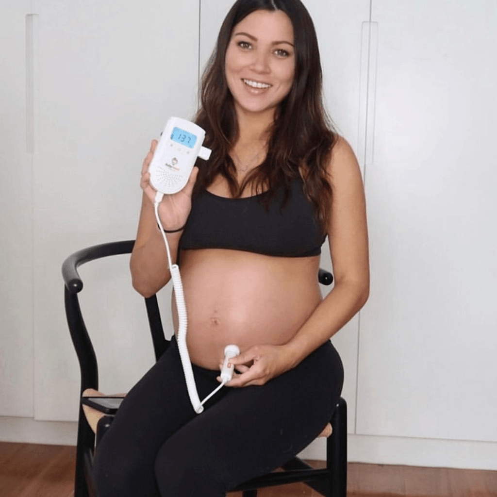 a smiling pregnant woman sitting on a chair holding the BabyHEart Standard Doppler listening to her baby's heart beat with 137 displayed on the doppler screen.